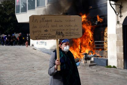 Una manifestante con una pancarta contra la reforma de las pensiones decretada por Macron pasa junto a la entrada en llamas de una oficina del banco BNP Paribas en Nantes, este martes. 